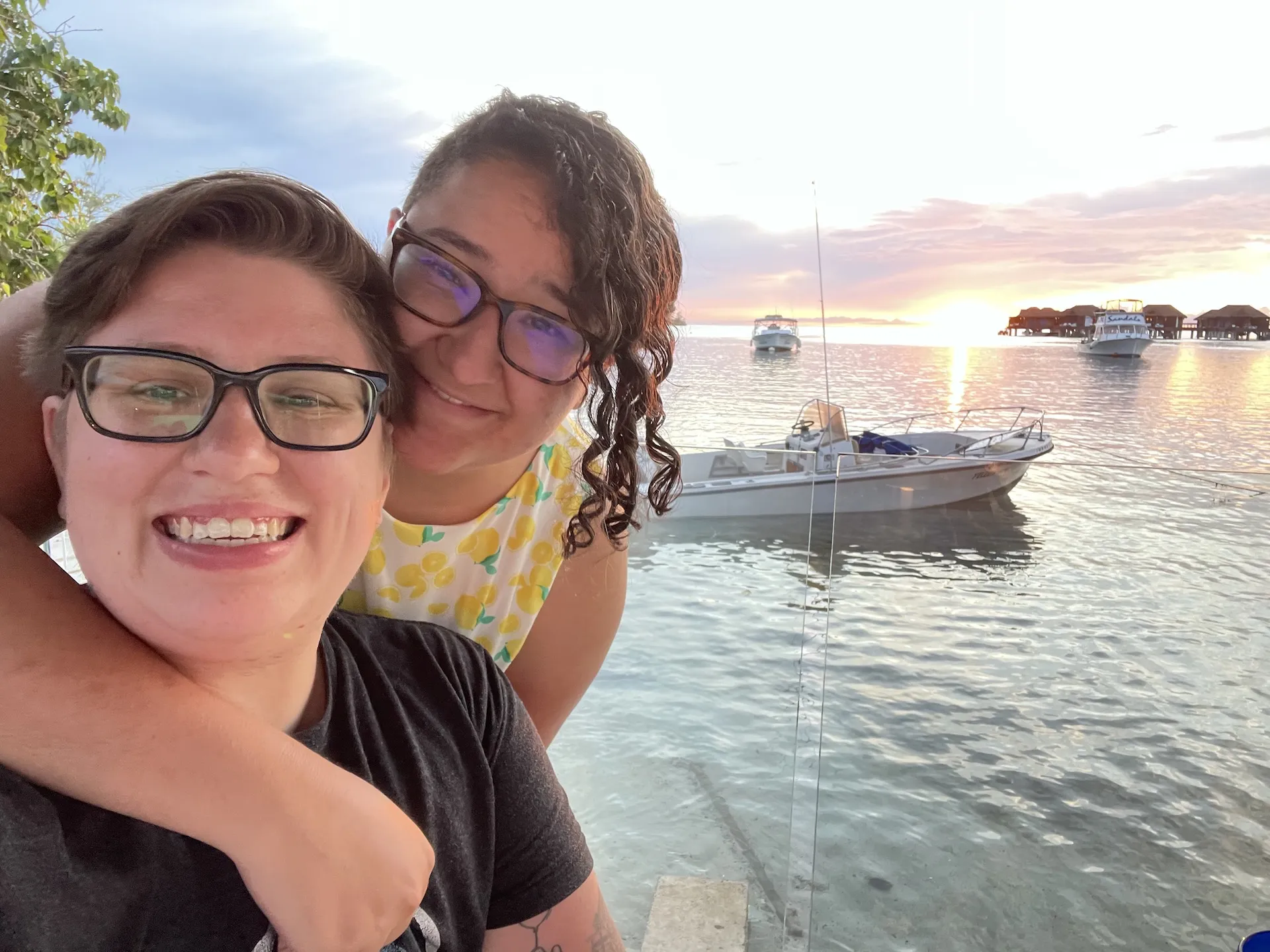 Andy a human with light brown short hair and glasses is smiling. They are wearing a dark gray t-shirt. Behind them is Ash who has black curly hair and glasses. They have their arm around Andy. In the background is a sunset on a bay with boats.