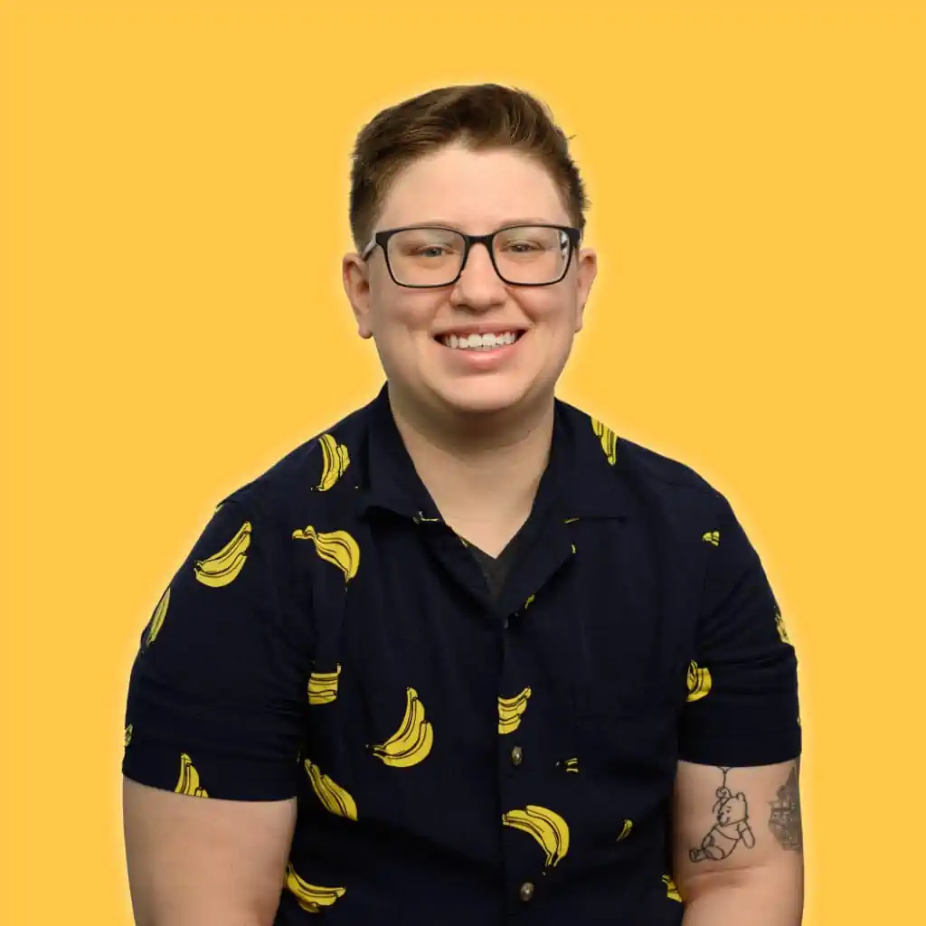 Andy, a human with short light brown hair and black glasses is smiling. They are wearing a button down shirt with a banana print. There are two black tattoos peeking out from the left sleeve. One is Pooh Bear floating with a balloon, the other is an architectural house from UP.