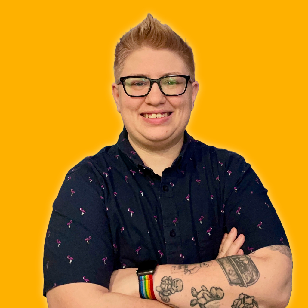 Andy is a human with short blonde hair and black glasses who is smiling. They are wearing a button-down shirt with a flamingo print. Their arms are crossed, exposing a sleeve of nerdy tattoos.